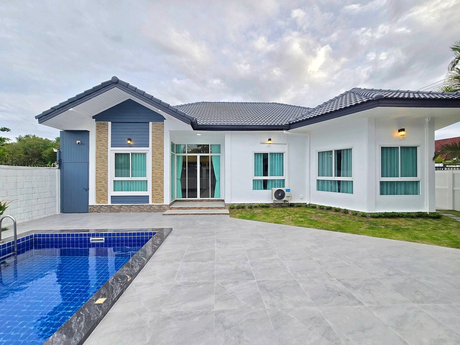 Mabprachan_Pool_Villa_Pattaya_Dlh571_18 Modern white house with blue accents and a blue-tiled pool in the foreground under a cloudy sky.