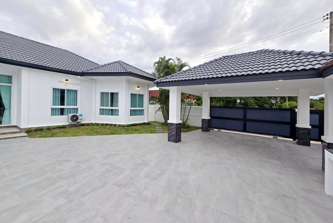 White modern house exterior with a gray tiled roof, large windows, and a covered carport/driveway area under a cloudy sky.