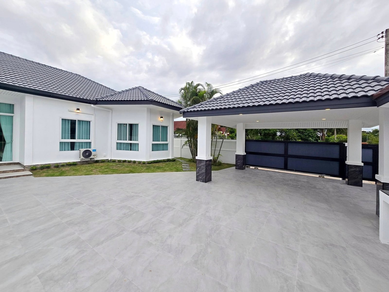 Mabprachan_Pool_Villa_Pattaya_Dlh571_19 White modern house exterior with a gray tiled roof, large windows, and a covered carport/driveway area under a cloudy sky.