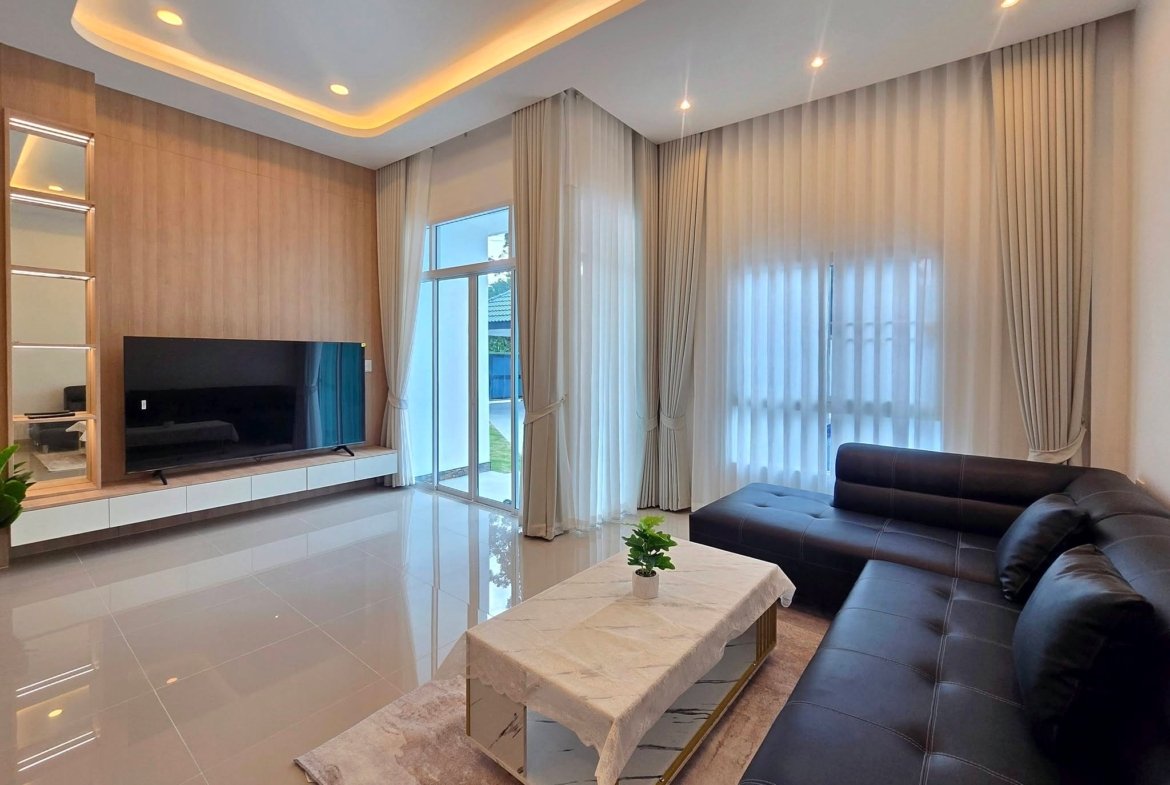 Modern living room with a black leather sectional, marble coffee table, and wall-mounted TV on a beige wood-paneled wall, next to large windows with sheer curtains.