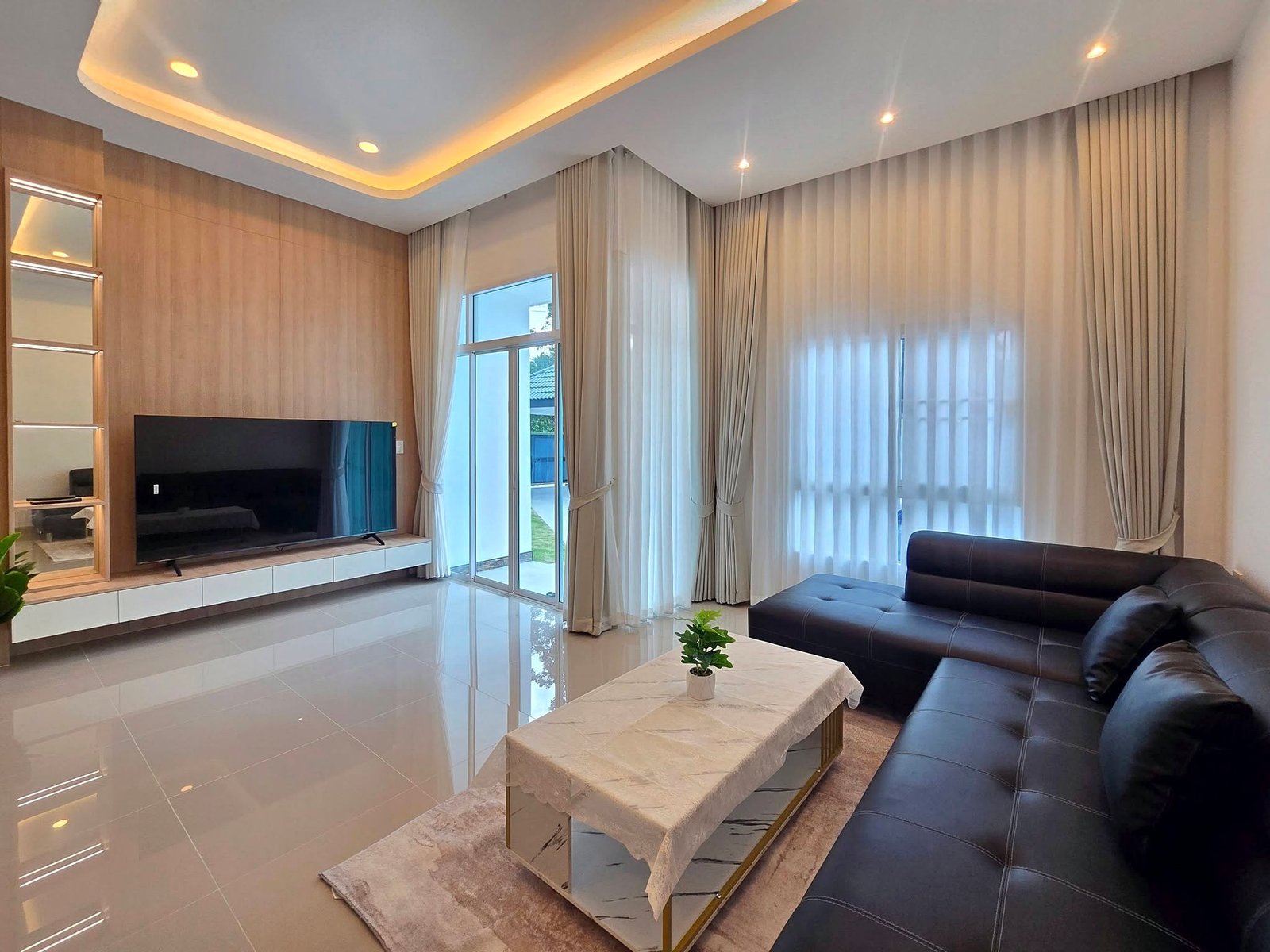 Mabprachan_Pool_Villa_Pattaya_Dlh571_3 Modern living room with a black leather sectional, marble coffee table, and wall-mounted TV on a beige wood-paneled wall, next to large windows with sheer curtains.