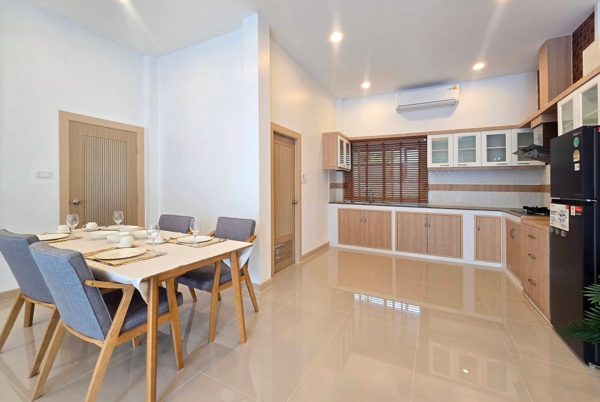 Bright open-plan dining and kitchen area with a wooden table set for six, grey chairs, white cabinetry, and wooden blinds.
