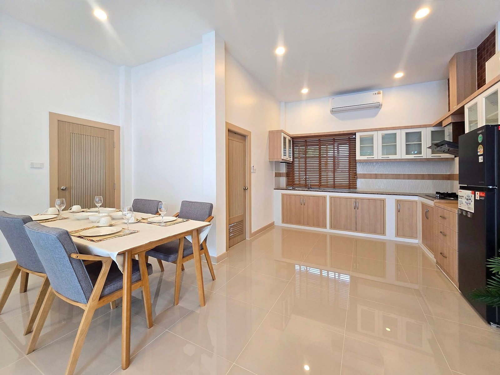 Mabprachan_Pool_Villa_Pattaya_Dlh571_4 Bright open-plan dining and kitchen area with a wooden table set for six, grey chairs, white cabinetry, and wooden blinds.