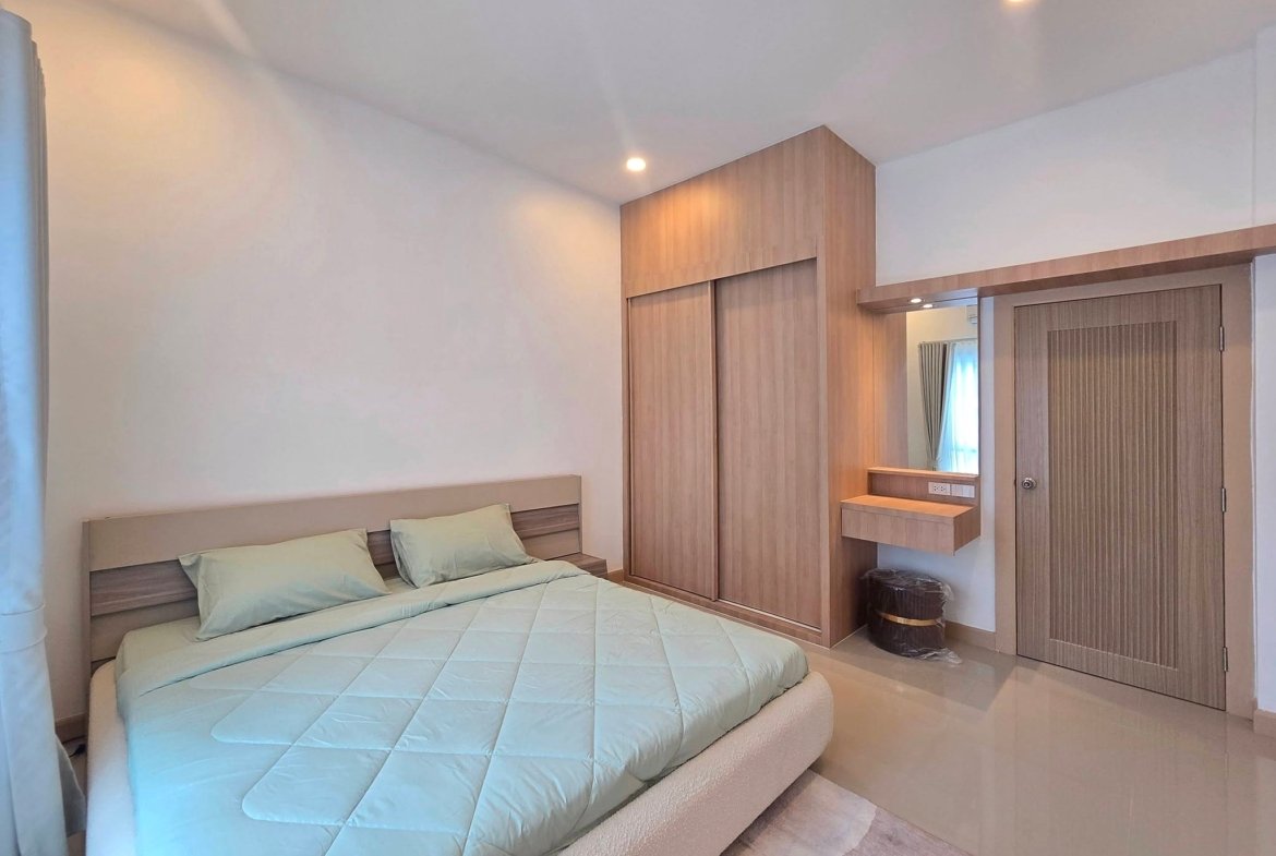 Bedroom with a large bed in pale blue bedding, and built-in wooden wardrobe and vanity along the right wall, lit by ceiling lights.