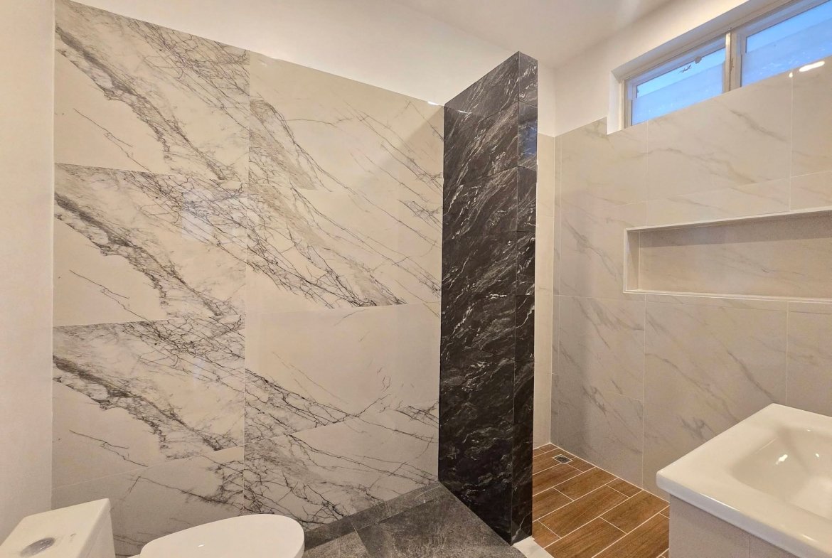 Contemporary bathroom with white marble walls, a dark marble pillar, and a wooden-look floor; toilet and bidet pair in the foreground and a sink near a window.