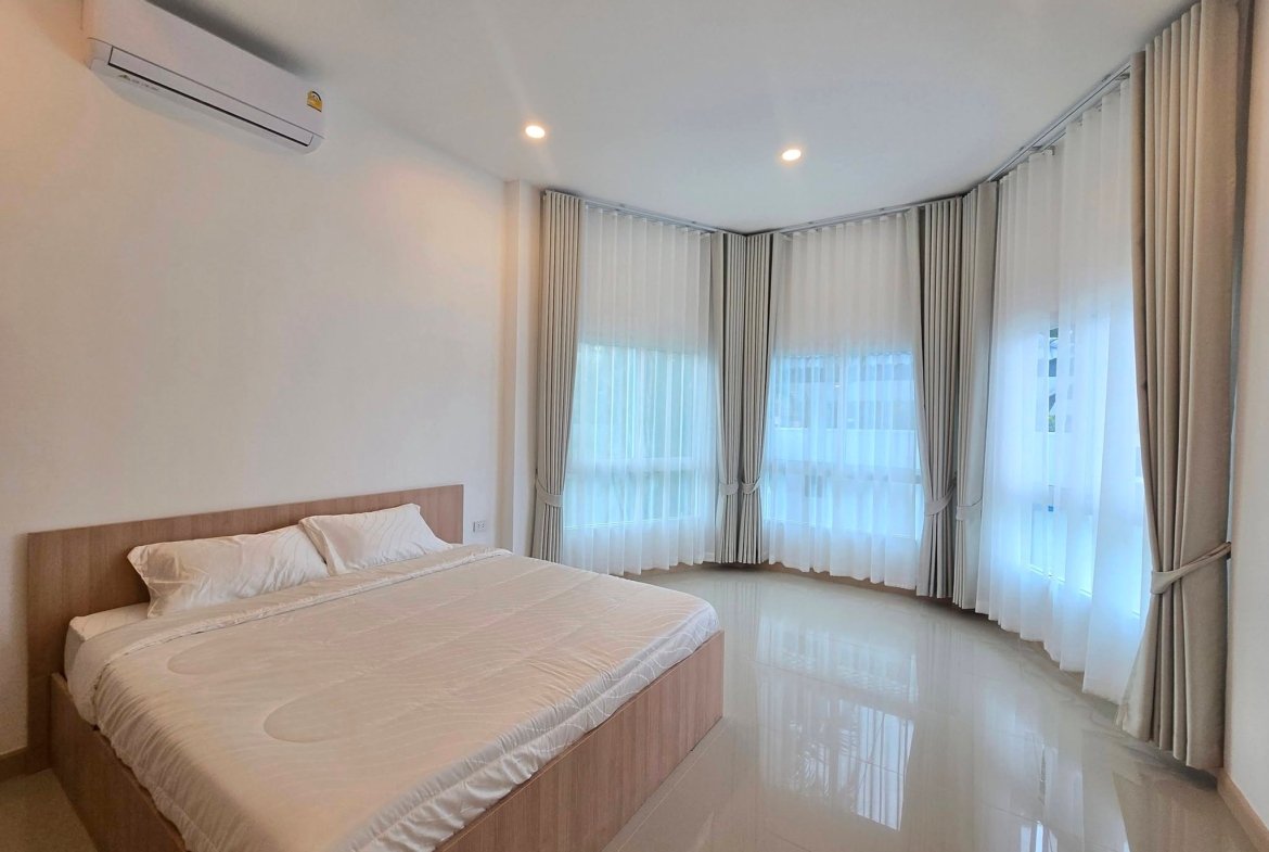 Twin-bed hotel room with white linens, wooden headboard, and glossy tiled floor; large windows with sheer and thick curtains along curved wall.