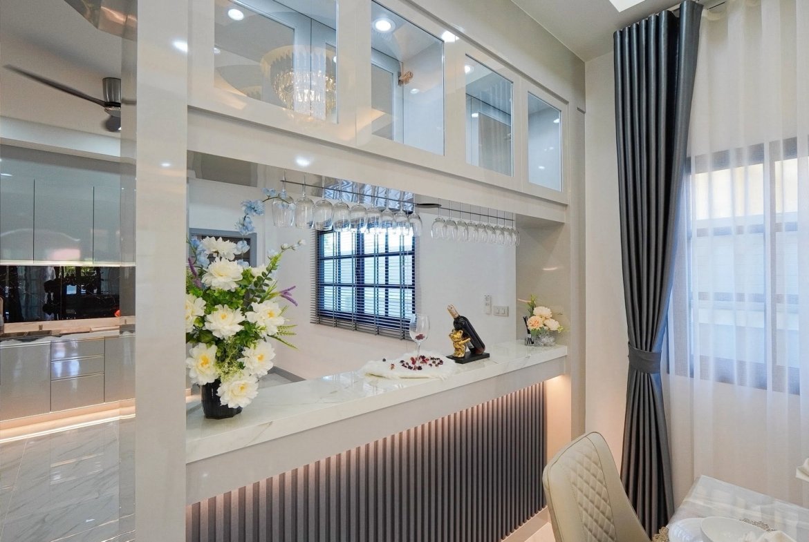 Modern kitchen/bar with wine glass rack, hanging glasses, and marble counter beneath white cabinets. Flowers and decorative items add elegance.