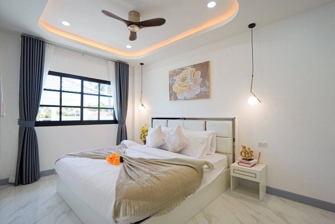 Modern bedroom with white bed, beige throw, orange flowers, and a curved, illuminated ceiling panel at nightfall while a ceiling fan spins overhead.