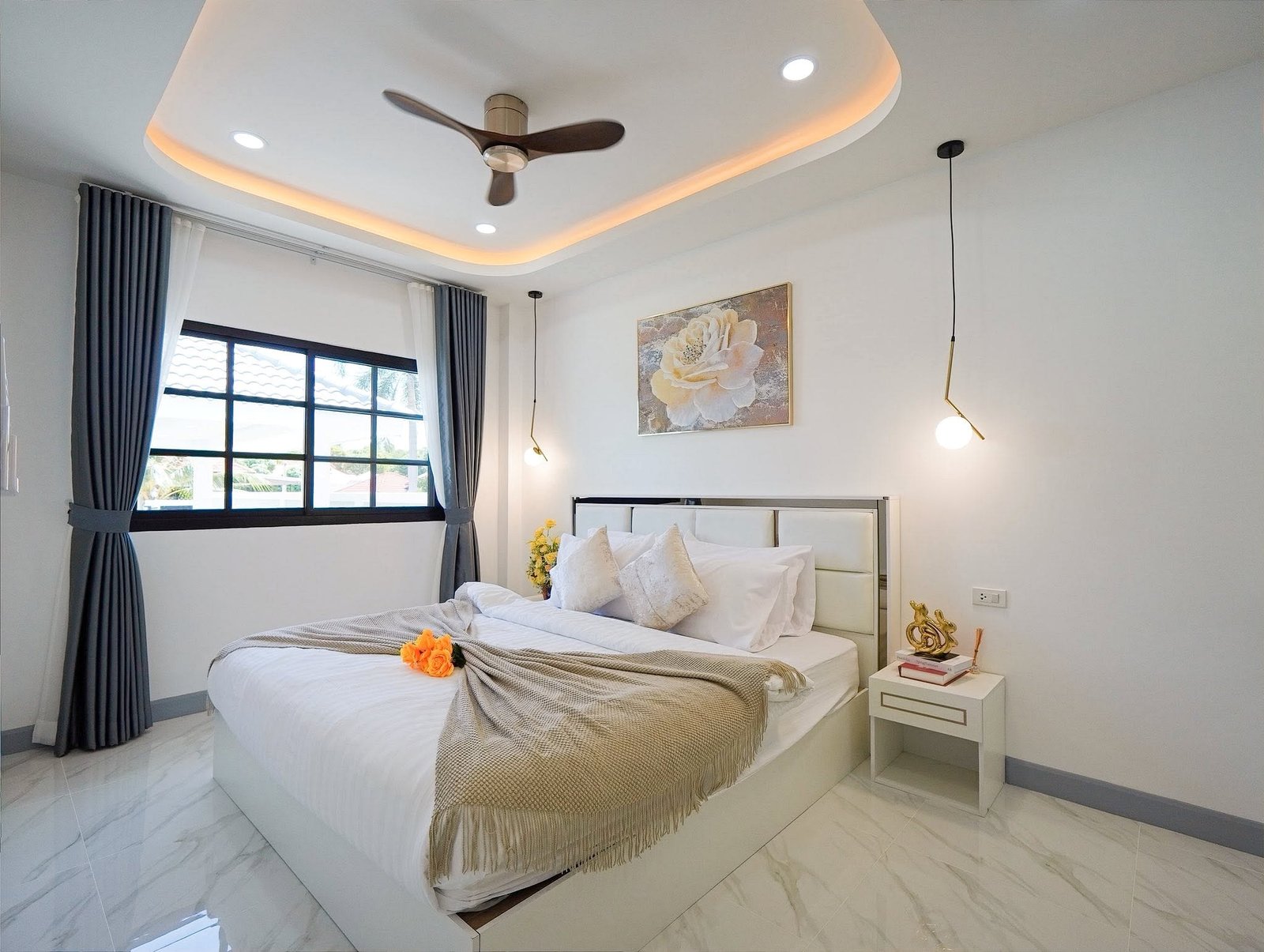 Mabprachan_Village_Pattaya_Dlh564_19 Modern bedroom with white bed, beige throw, orange flowers, and a curved, illuminated ceiling panel at nightfall while a ceiling fan spins overhead.
