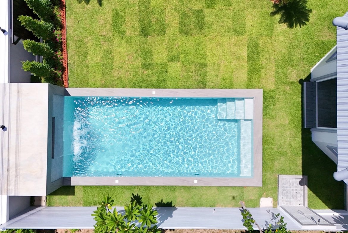 Aerial top-down view of a rectangular turquoise swimming pool surrounded by light-gray coping, green lawn, and white buildings with palm trees casting shadows.
