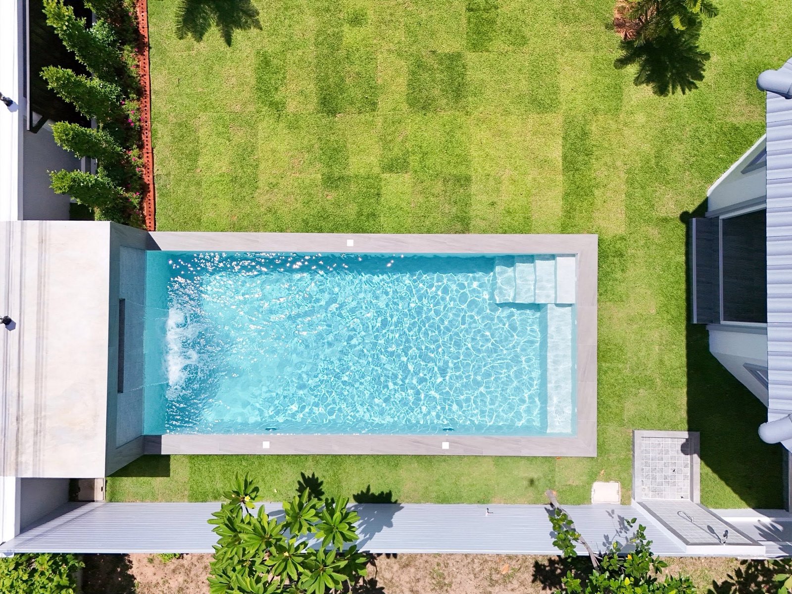 Mabprachan_Village_Pattaya_Dlh564_2 Aerial top-down view of a rectangular turquoise swimming pool surrounded by light-gray coping, green lawn, and white buildings with palm trees casting shadows.
