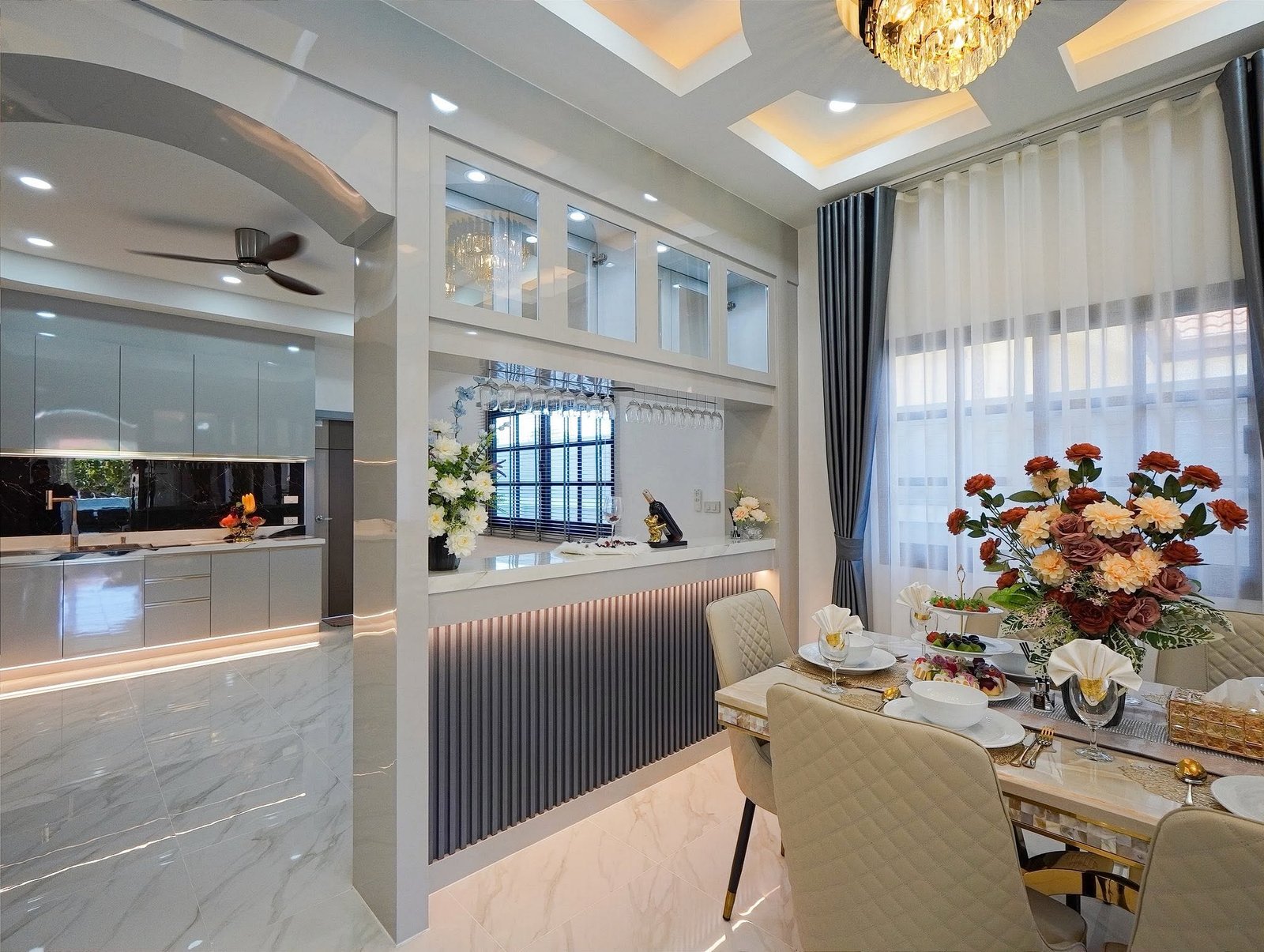 Mabprachan_Village_Pattaya_Dlh564_26 Open-concept kitchen and dining area with white cabinetry, glass-front uppers, hanging wine glasses, and a large floral centerpiece.