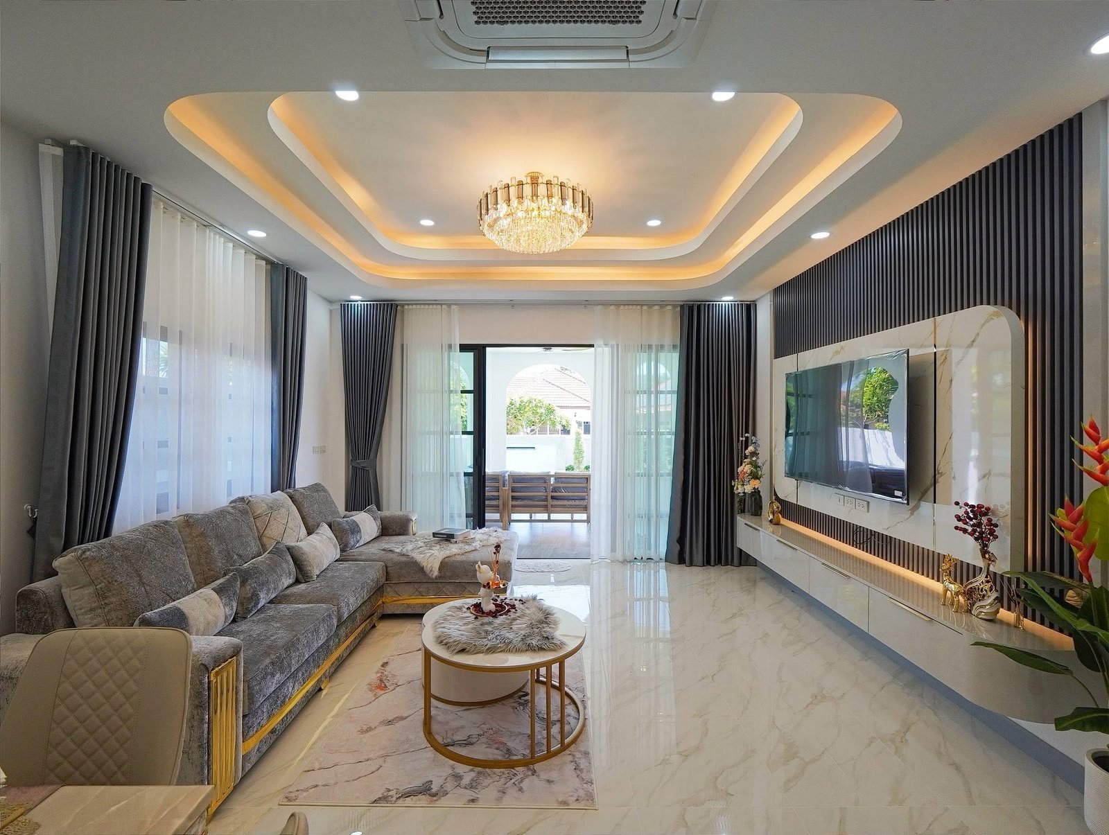 Mabprachan_Village_Pattaya_Dlh564_3 Luxurious living room with a crystal chandelier, layered ceiling arches, and a gray sectional sofa.