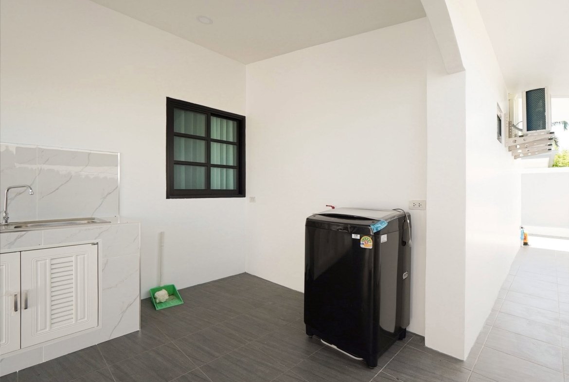 Outdoor laundry area with a black washing machine beside white cabinets and a marble-topped sink area; grey tiled floor and a dark-framed window on the wall.