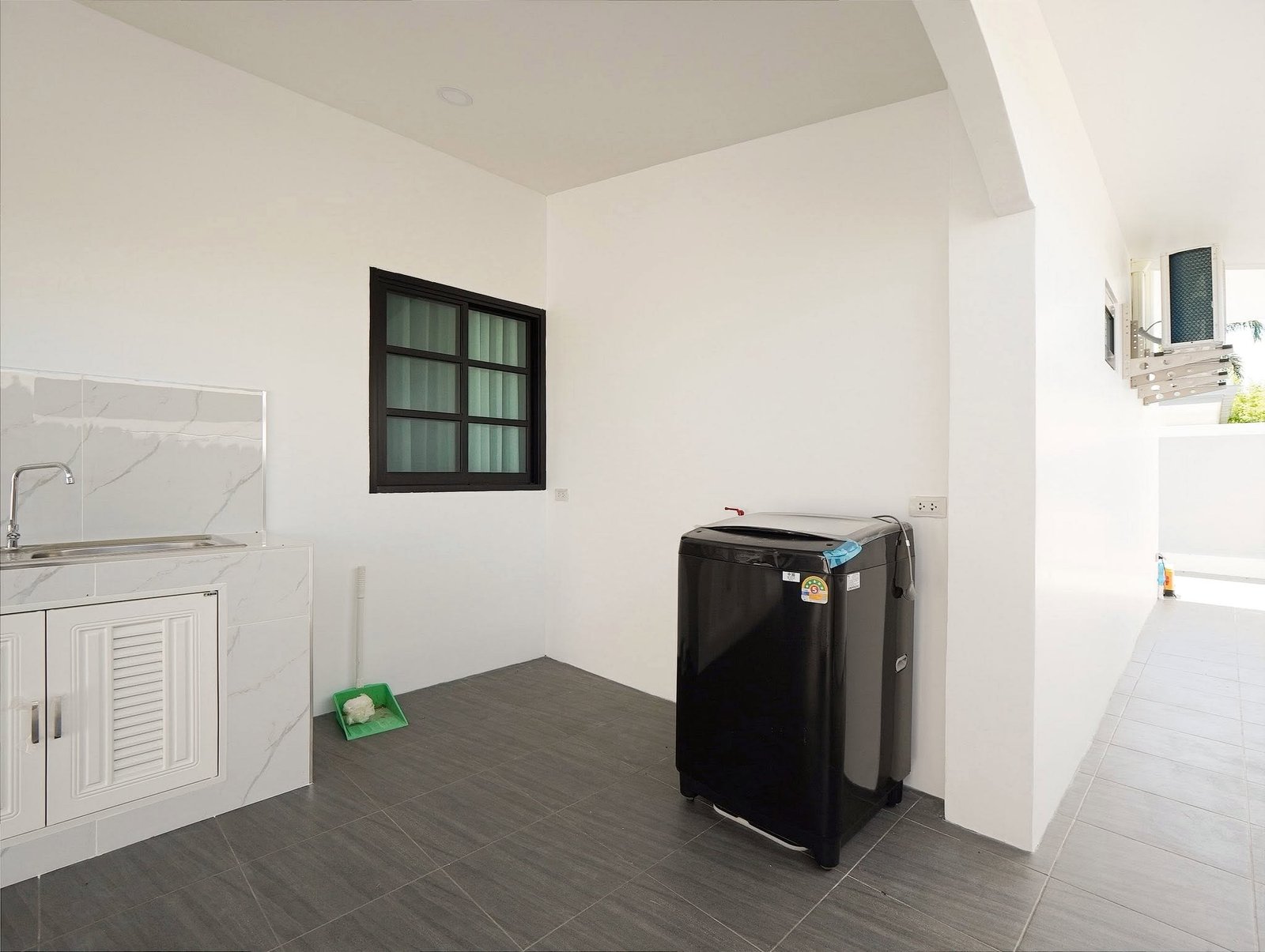 Mabprachan_Village_Pattaya_Dlh564_40 Outdoor laundry area with a black washing machine beside white cabinets and a marble-topped sink area; grey tiled floor and a dark-framed window on the wall.