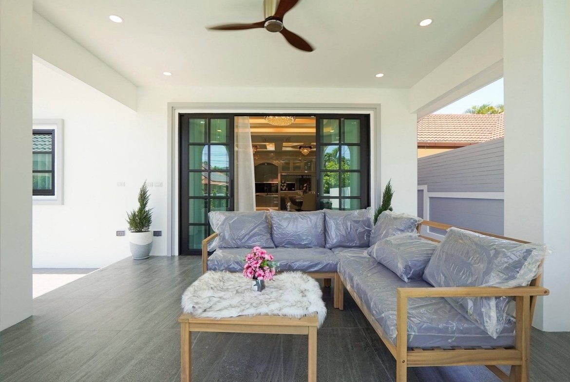 Covered outdoor seating area with wooden-frame sofa and gray cushions, coffee table with pink flowers, tiled floor, and ceiling fan above.