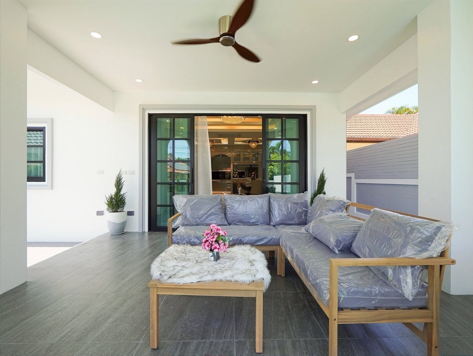 Mabprachan_Village_Pattaya_Dlh564_41 Covered outdoor seating area with wooden-frame sofa and gray cushions, coffee table with pink flowers, tiled floor, and ceiling fan above.