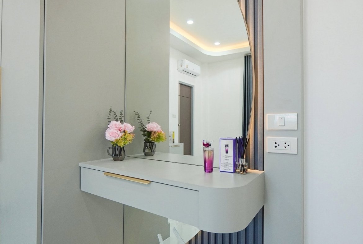 Modern vanity area with gray cabinetry, a curved mirror, and a white countertop holding pink flowers in glass vases and purple cosmetic packaging nearby.
