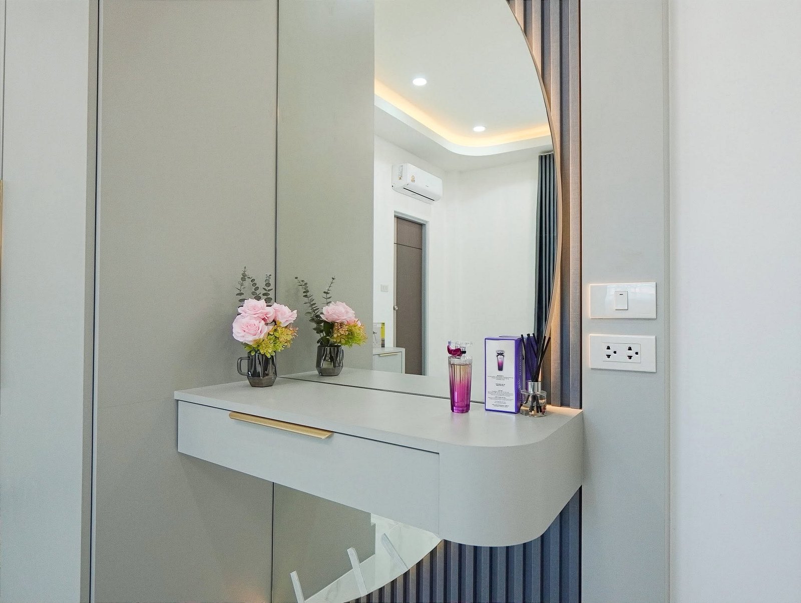 Mabprachan_Village_Pattaya_Dlh564_9 Modern vanity area with gray cabinetry, a curved mirror, and a white countertop holding pink flowers in glass vases and purple cosmetic packaging nearby.