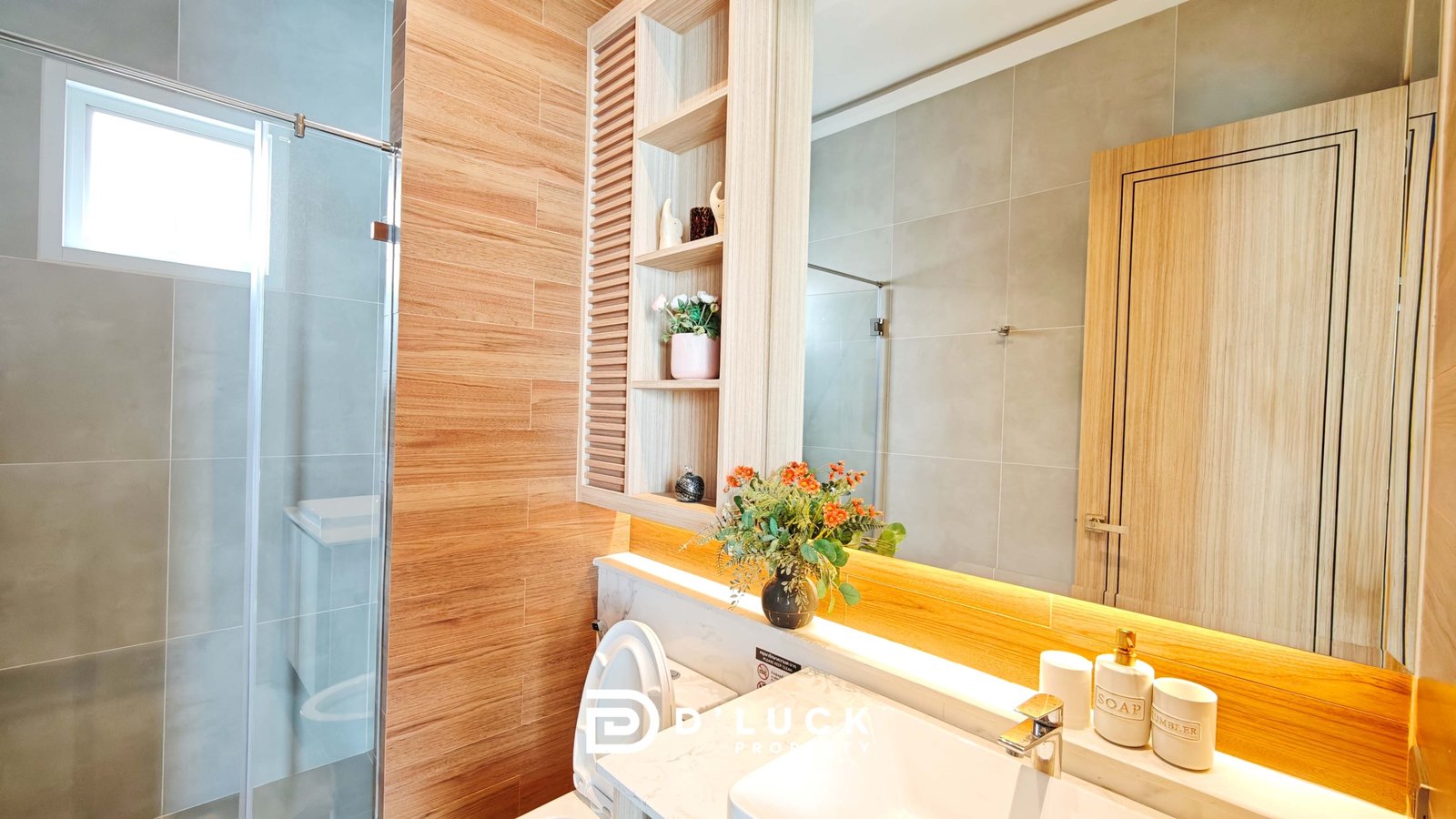 Modern bathroom with glass shower door, tan tiles, and a wooden shelving unit beside the sink