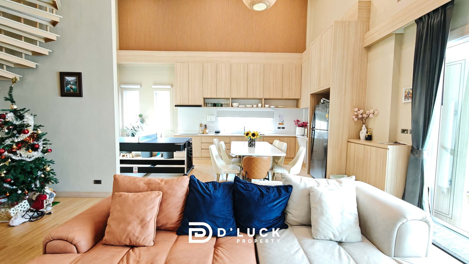 Open-plan living area with a Christmas tree, peach and navy cushions on a beige sofa, and light wood kitchen cabinets.