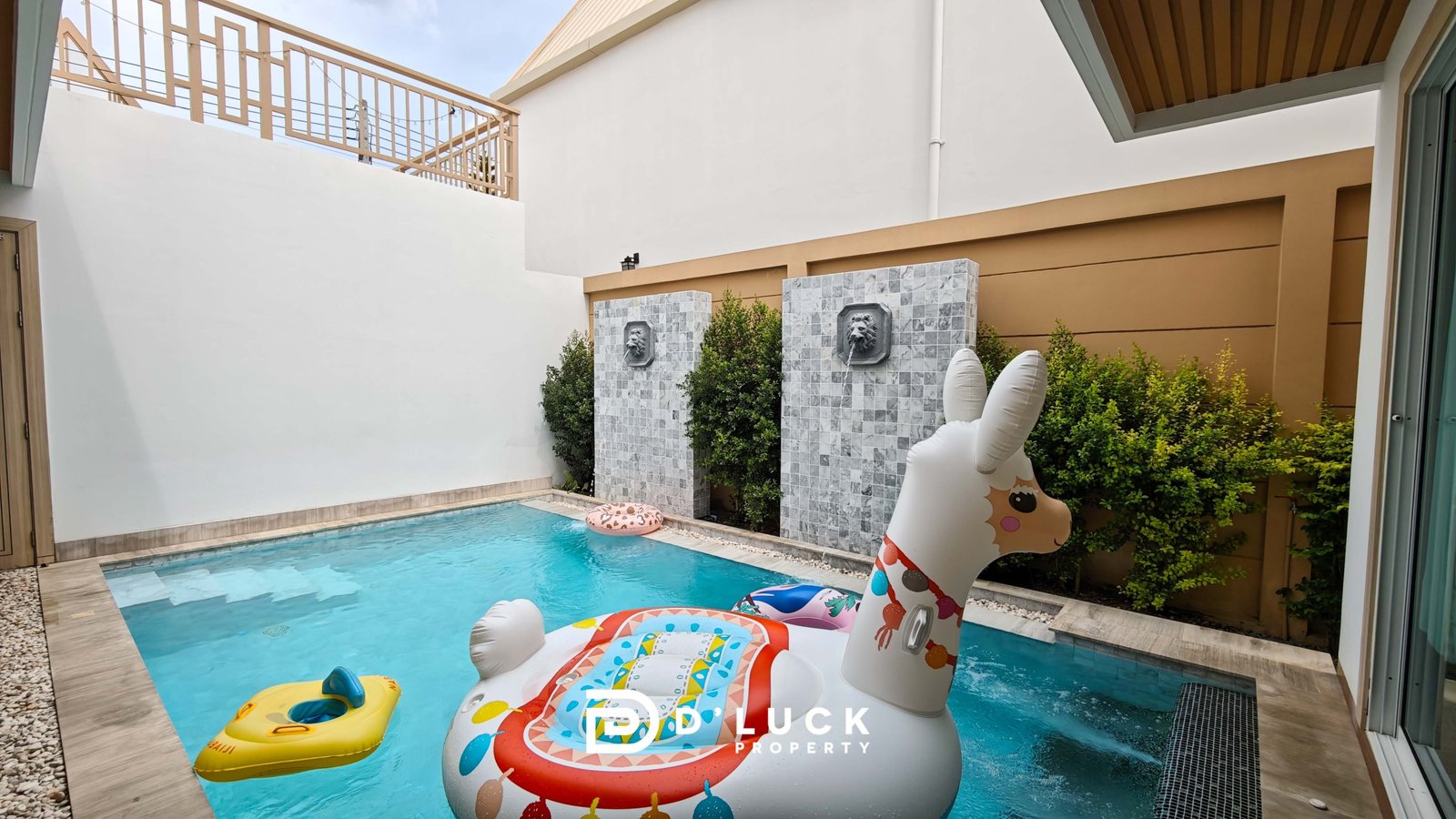 Pool in a courtyard with inflatable unicorn and yellow raft on blue water; decorative lion-head fountains on tiled stone columns and greenery nearby.