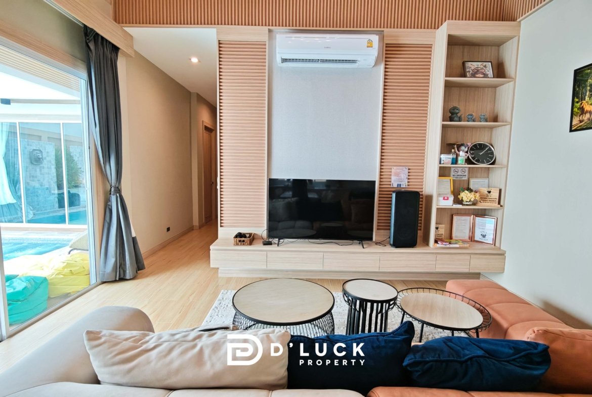 Modern living room with a built-in wooden media wall, wall-mounted TV, open shelving, and orange seating; sliding glass doors lead to a pool area outside. An air conditioner sits above the TV.