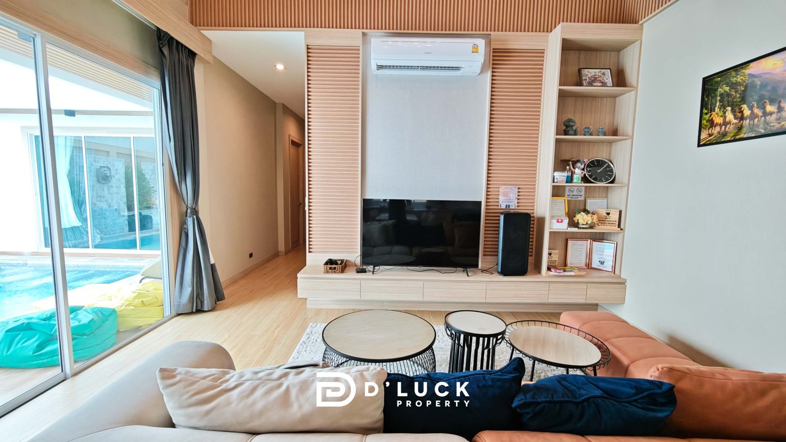 Modern living room with a built-in wooden media wall, wall-mounted TV, open shelving, and orange seating; sliding glass doors lead to a pool area outside. An air conditioner sits above the TV.