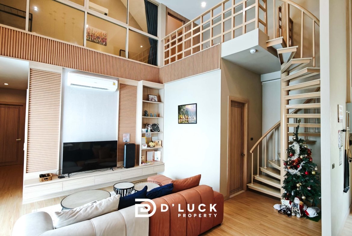 Living room with beige L-shaped sofa, round coffee tables, wall TV, built-in shelves, and a wooden staircase to the mezzanine level.
