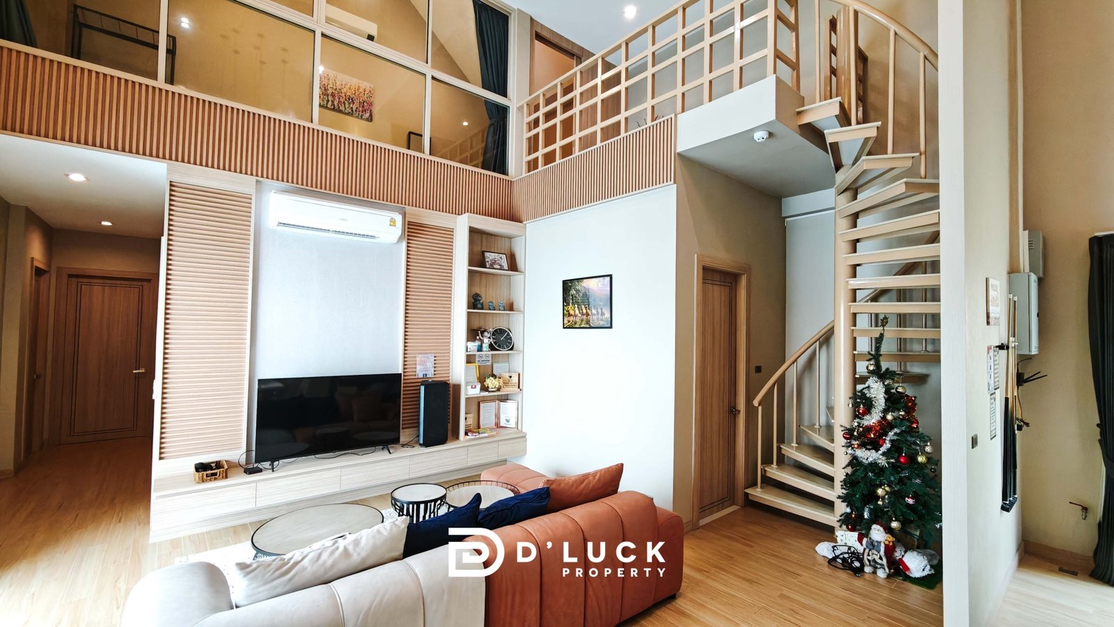 Living room with beige L-shaped sofa, round coffee tables, wall TV, built-in shelves, and a wooden staircase to the mezzanine level.