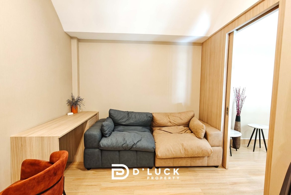 Cozy living room with beige walls, a two-tone sectional (gray and tan), a wooden desk along the left wall, and a sliding door on the right.