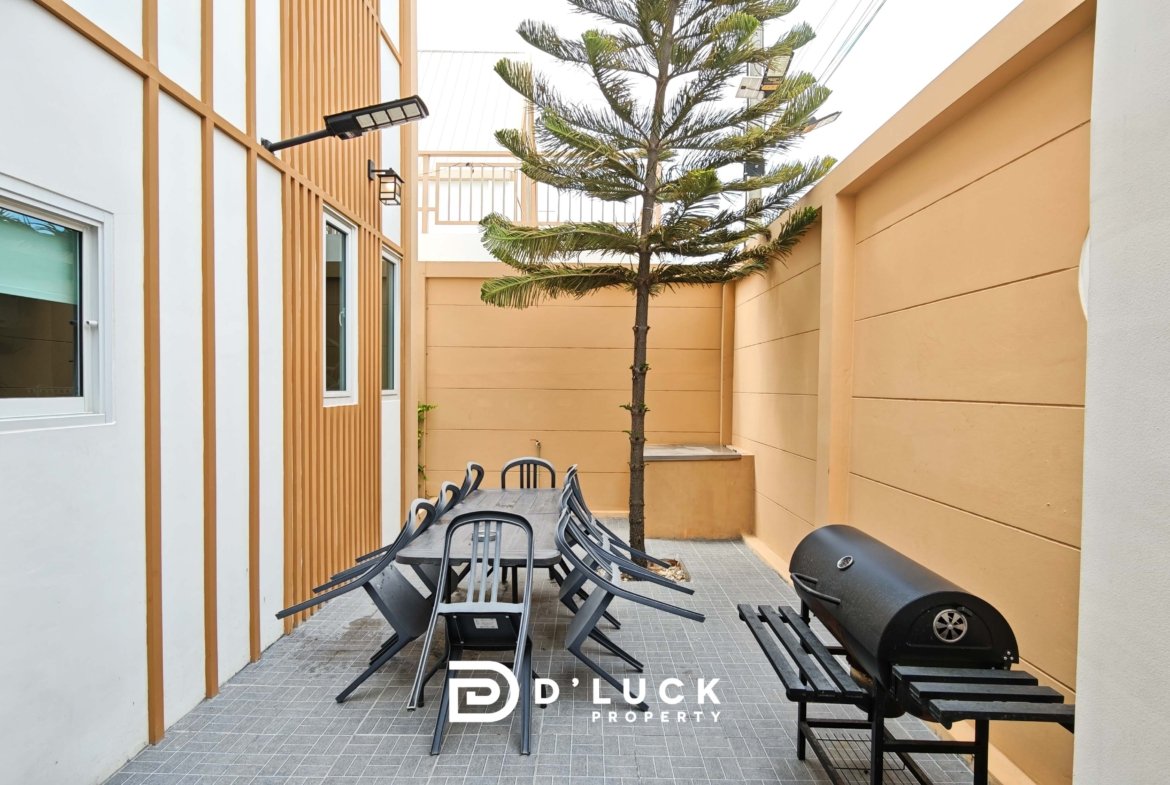 Outdoor courtyard with a metal table and stacked black chairs, flanked by orange-brown walls and white panels, and a tall tree in the center.