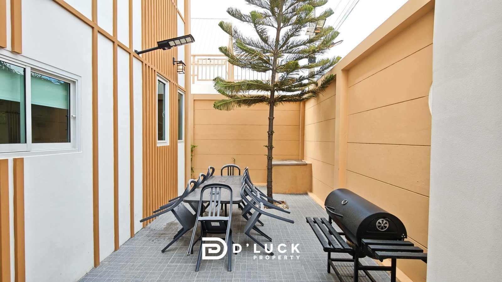 Outdoor courtyard with a metal table and stacked black chairs, flanked by orange-brown walls and white panels, and a tall tree in the center.