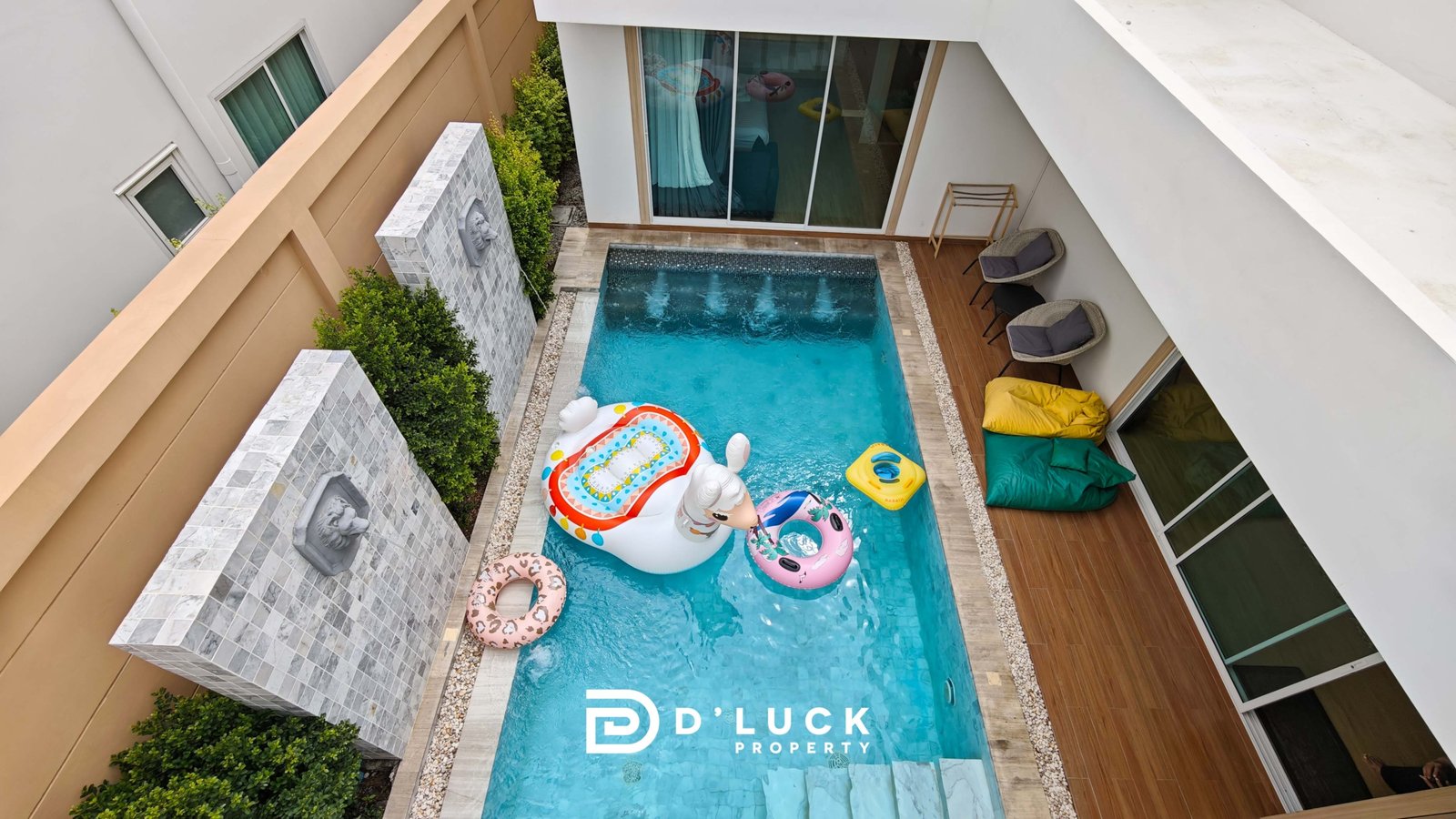 Top-down view of a rectangular pool in a private courtyard, with colorful inflatable floats and a wooden deck along the sides.
