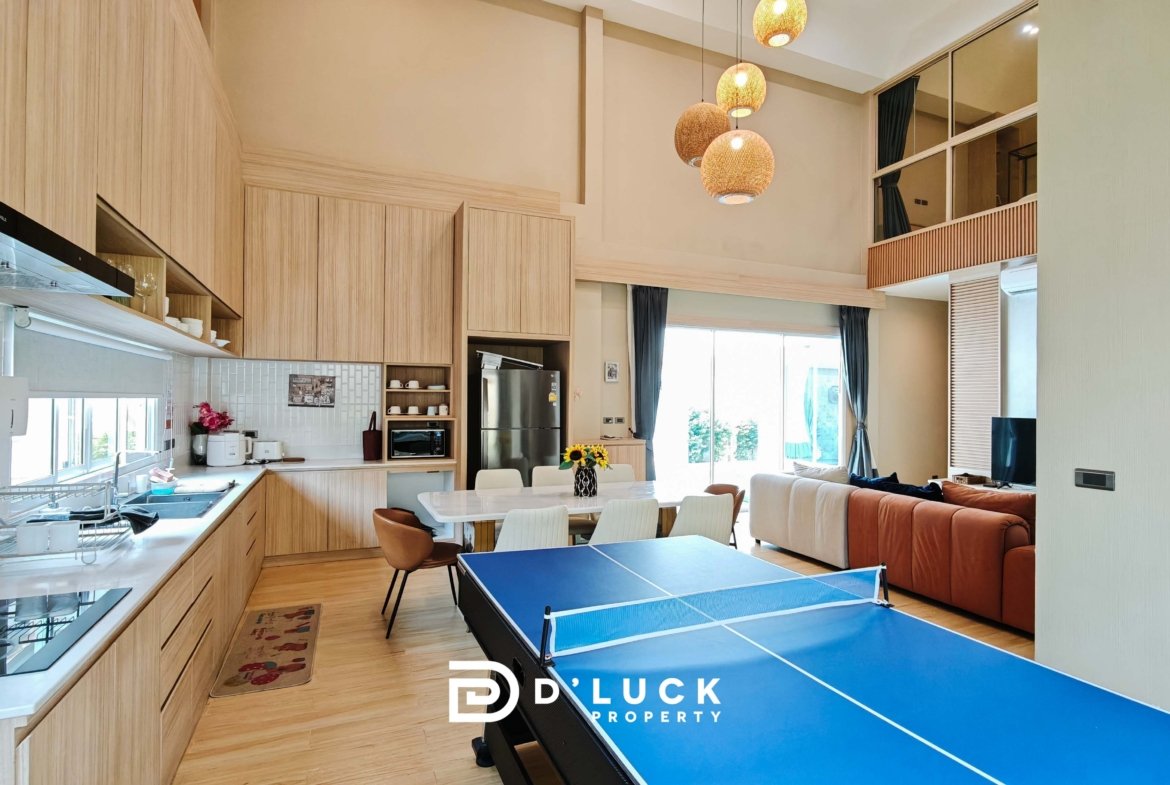 Open-plan kitchen and living area with light wood cabinets, wicker pendant lights, and a blue ping-pong table in the foreground, sofa in back.