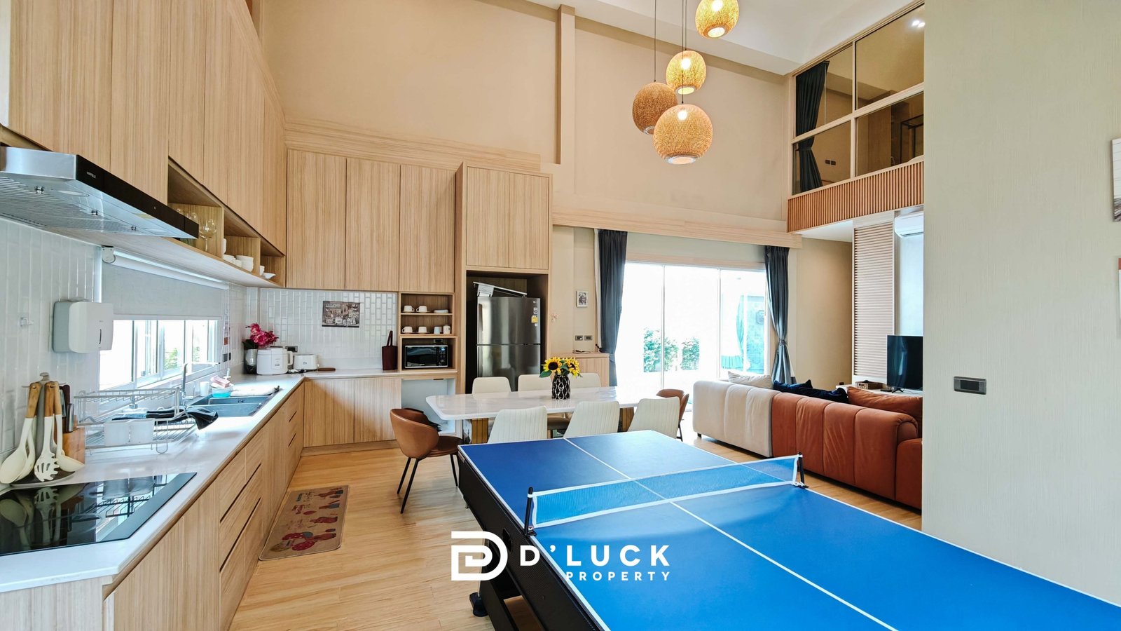 Open-plan kitchen and living area with light wood cabinets, wicker pendant lights, and a blue ping-pong table in the foreground, sofa in back.