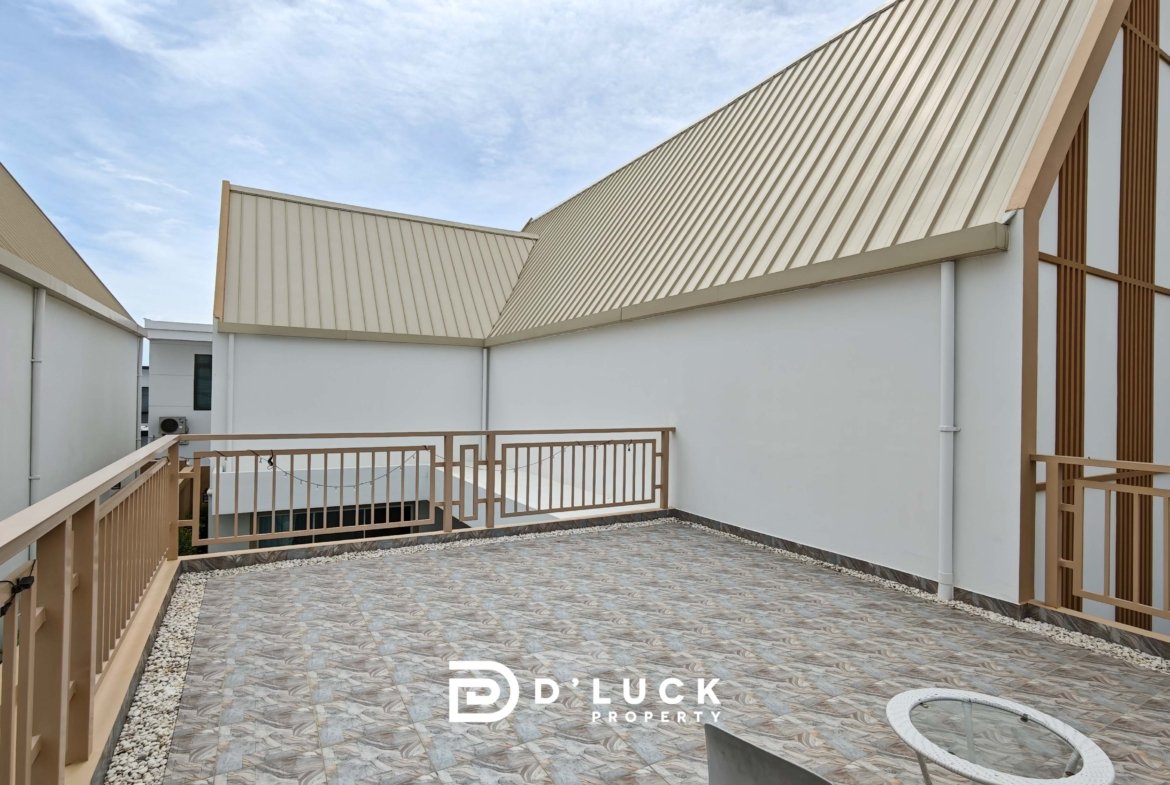 Rooftop terrace of a modern building with beige slanted metal roofs, white walls, and brown railings against a blue sky.