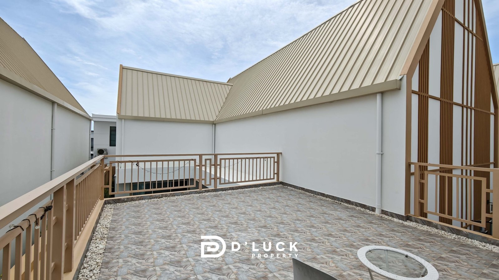 Rooftop terrace of a modern building with beige slanted metal roofs, white walls, and brown railings against a blue sky.