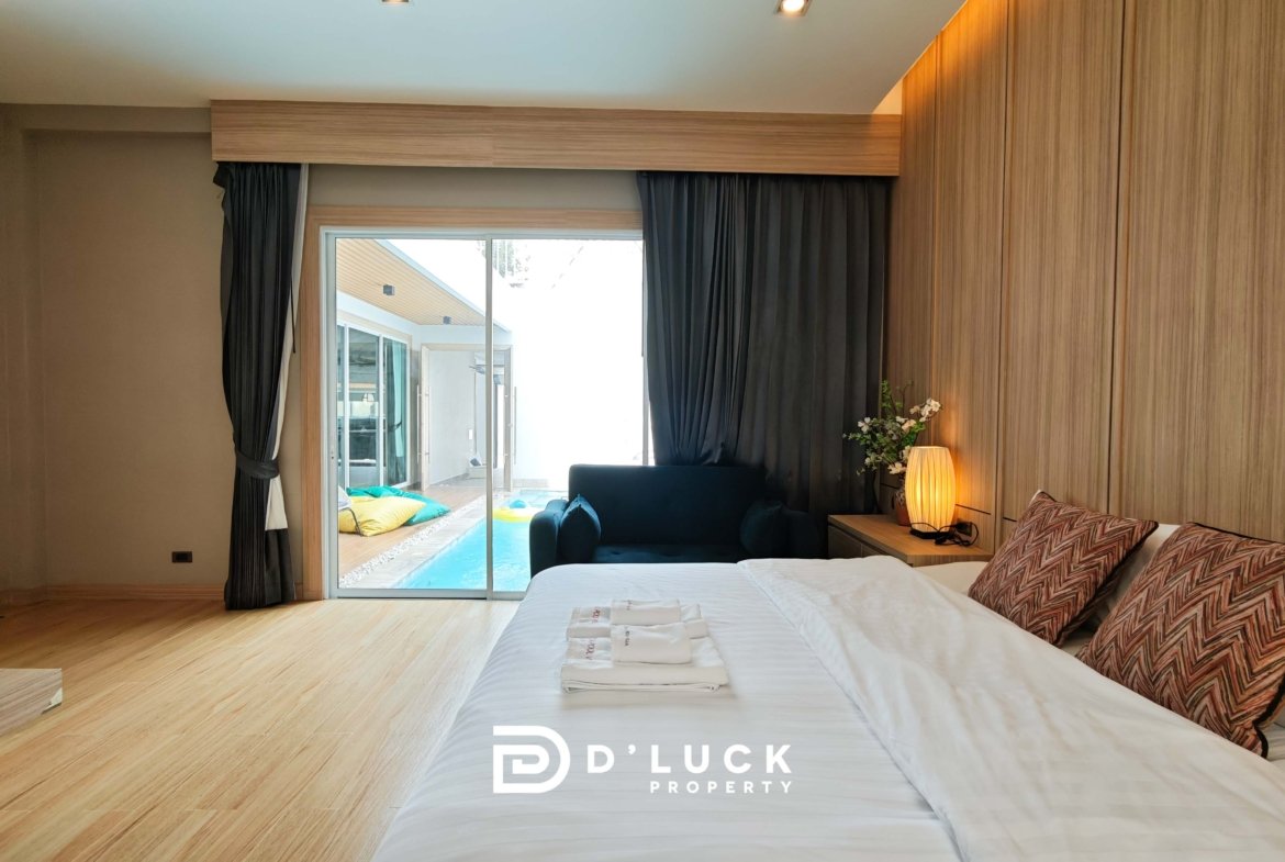 Modern bedroom with white bed, patterned pillows, wood-paneled wall, and sliding glass door to the pool patio outside.