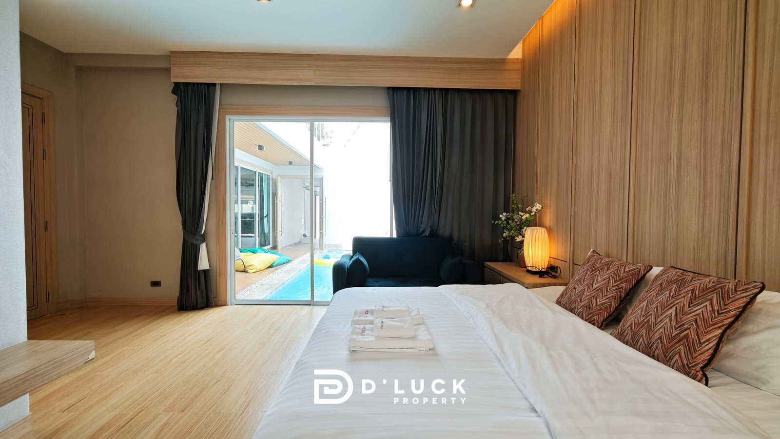 Modern bedroom with white bed, patterned pillows, wood-paneled wall, and sliding glass door to the pool patio outside.