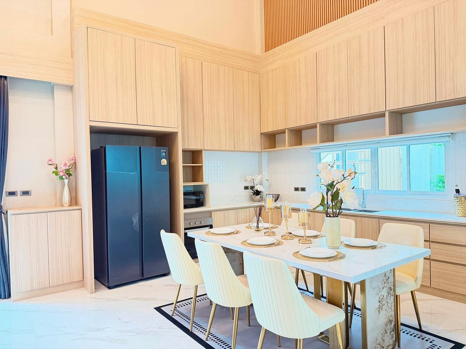 Narita_Villa_Pattaya_Rent_Rdl339_10 Bright open kitchen with light wood cabinets, a marble dining table set for six, gold candles, and white chairs; blue refrigerator on the left and flowers on the counter.
