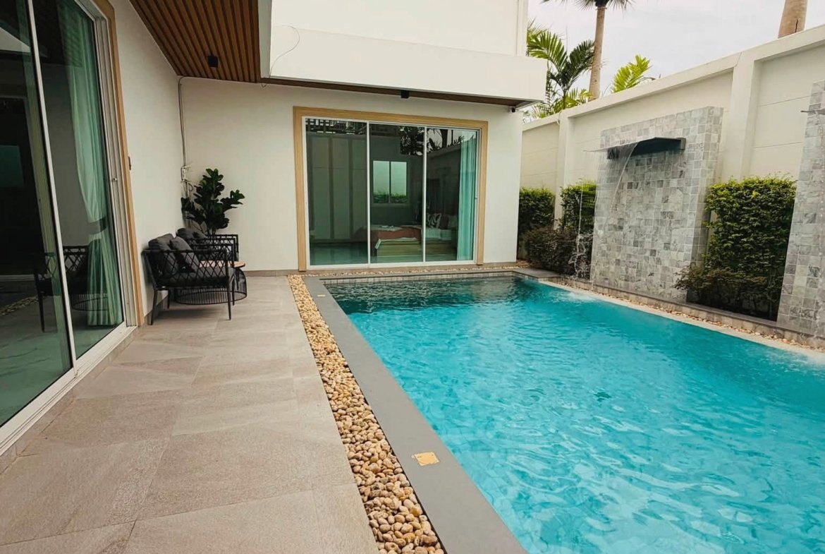 Modern backyard with a rectangular blue pool, beige stone deck, and a stone pebble border next to glass sliding doors.