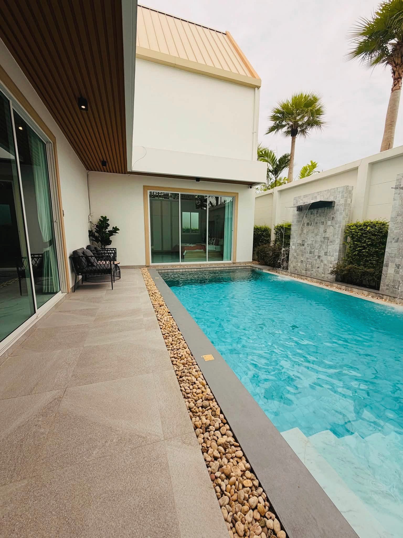 Narita_Villa_Pattaya_Rent_Rdl339_2 Modern backyard with a rectangular blue pool, beige stone deck, and a stone pebble border next to glass sliding doors.