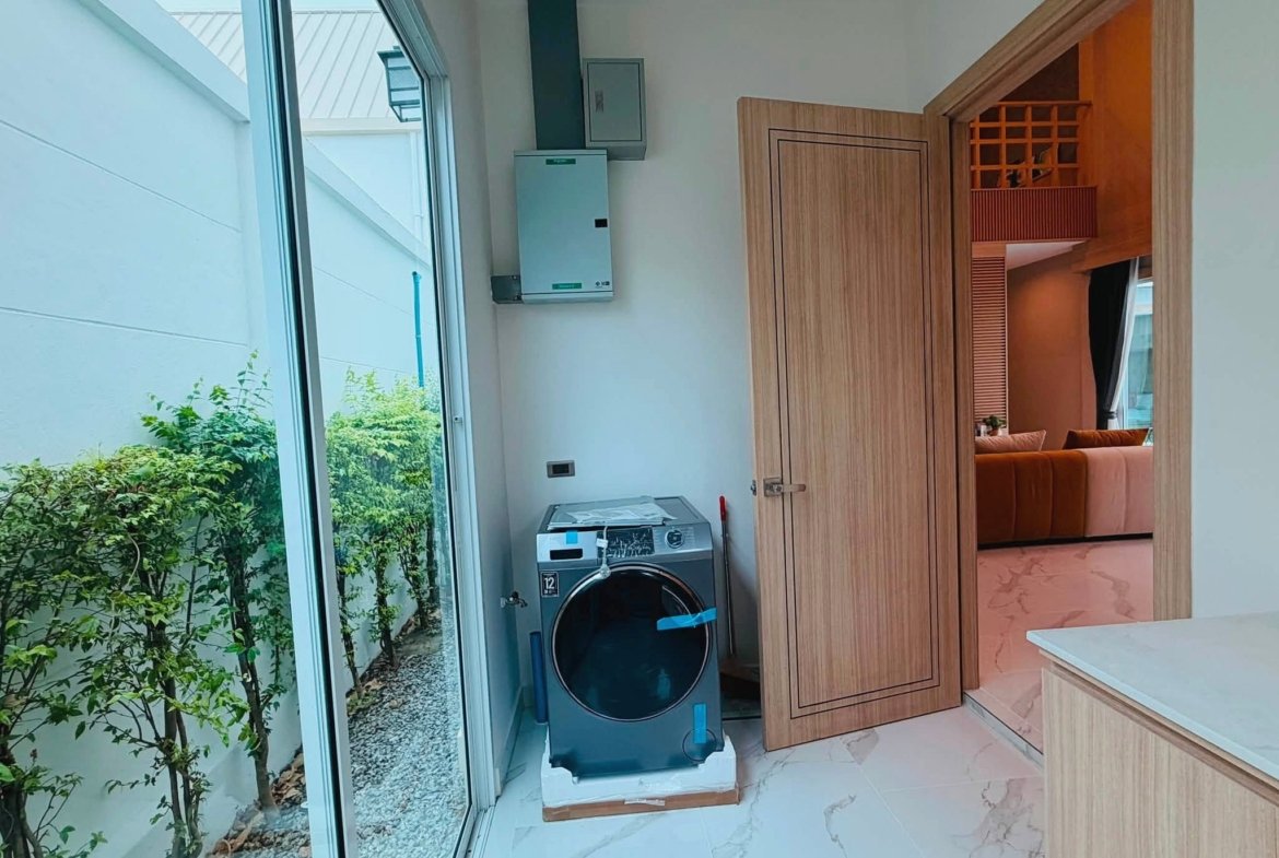 Laundry area with a stacked washer and dryer beside a wooden cabinet, glass door to a garden on the left.