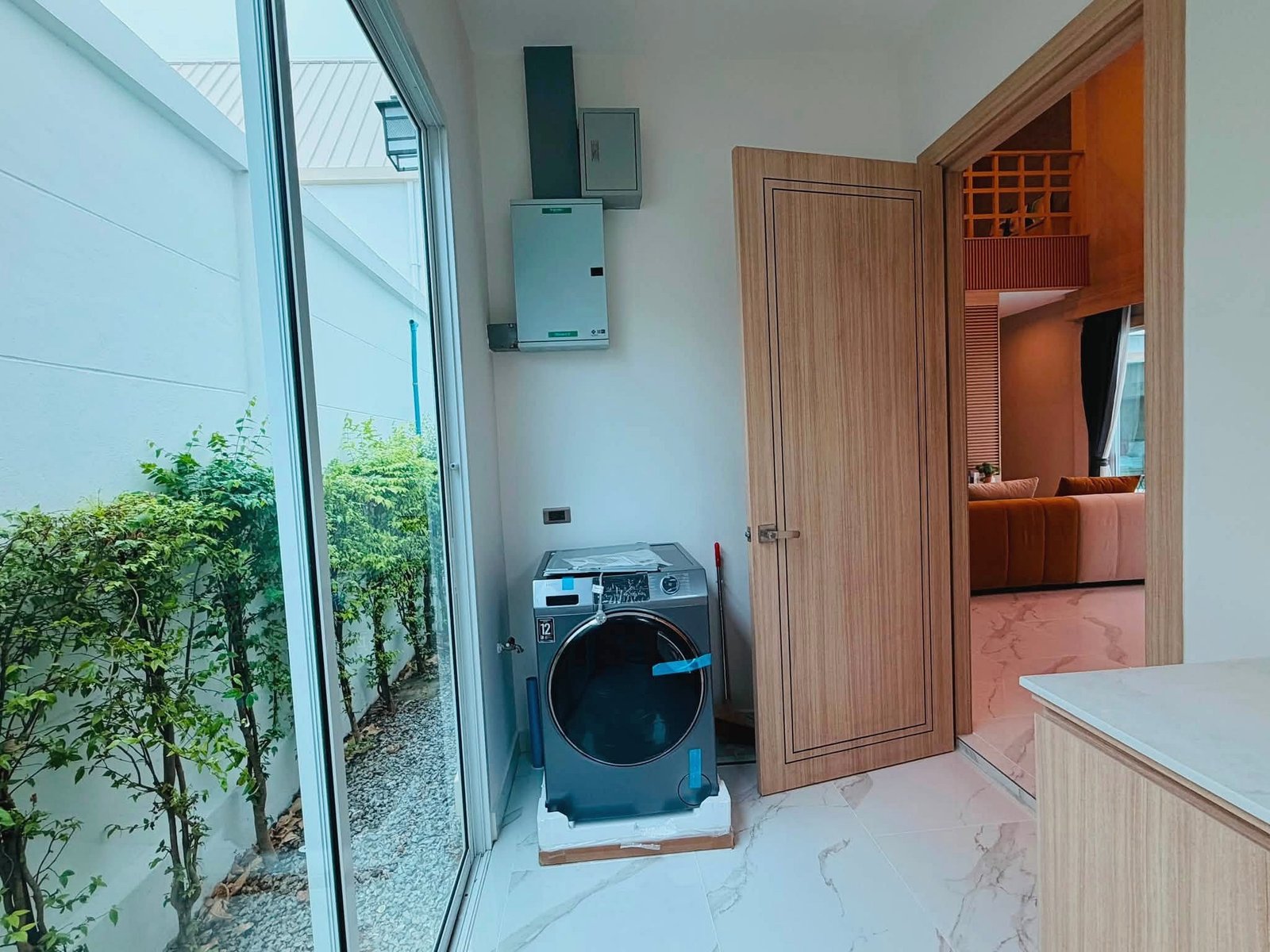 Narita_Villa_Pattaya_Rent_Rdl339_22 Laundry area with a stacked washer and dryer beside a wooden cabinet, glass door to a garden on the left.