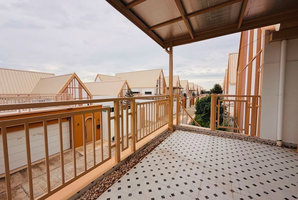 Outdoor walkway of a residential complex with beige metal railings, tiled floor, and stone edging, row of cream-colored houses with pitched roofs in the background under a cloudy sky.