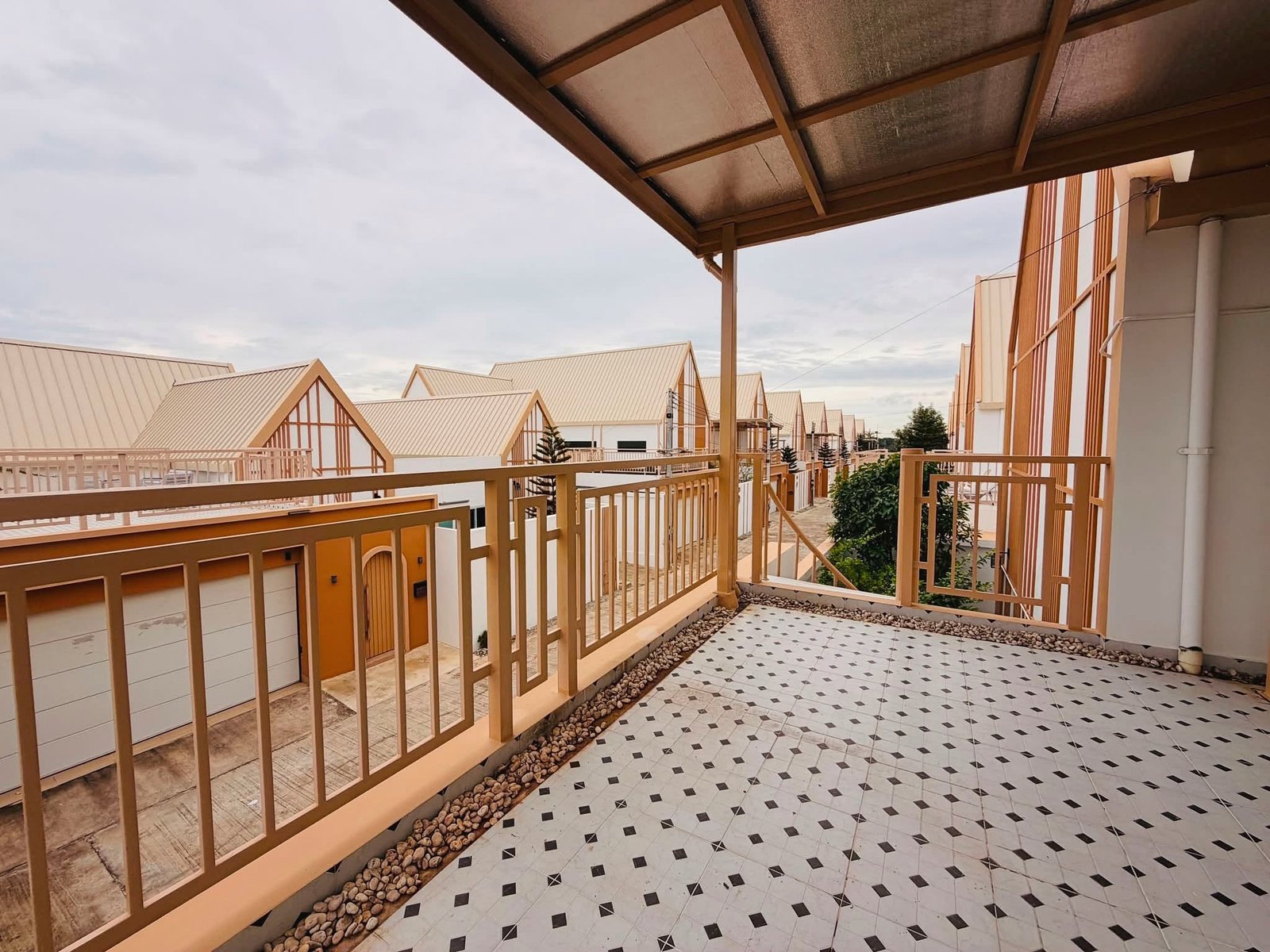Narita_Villa_Pattaya_Rent_Rdl339_24 Outdoor walkway of a residential complex with beige metal railings, tiled floor, and stone edging, row of cream-colored houses with pitched roofs in the background under a cloudy sky.