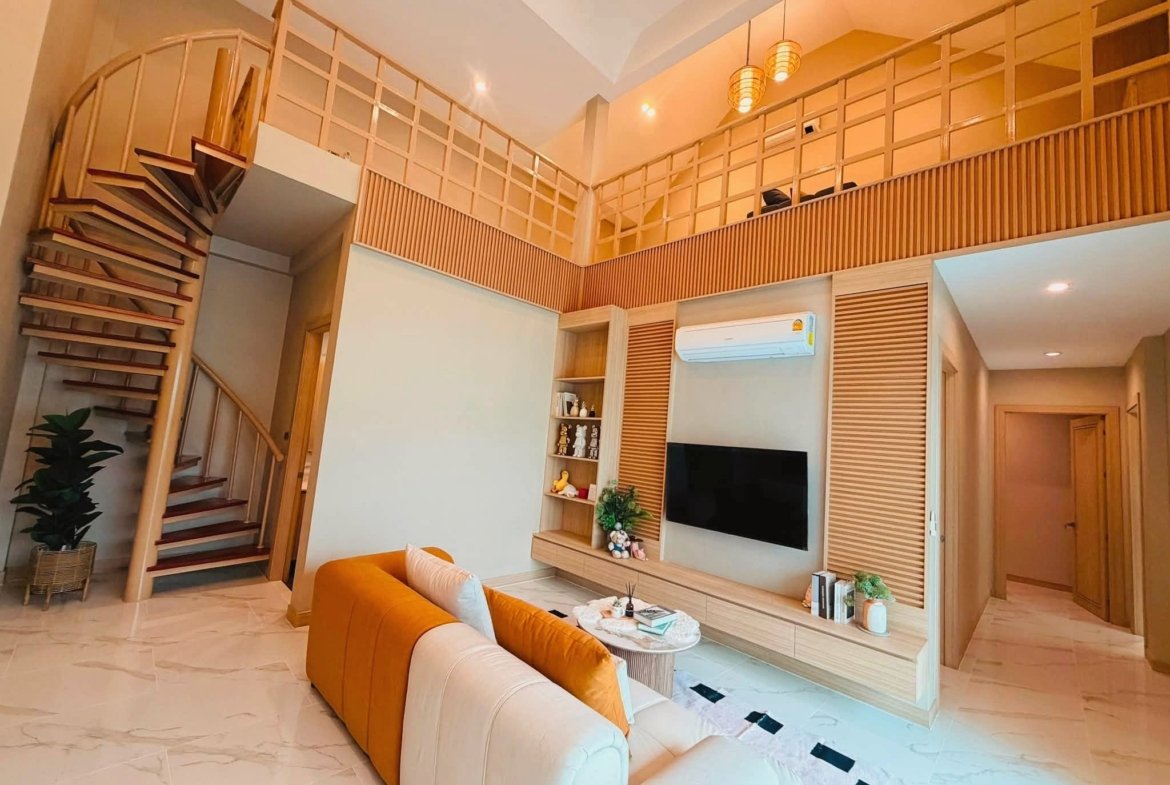 Open living room with orange and beige sectional, wall-mounted TV, and a wooden staircase leading to a mezzanine on the left.