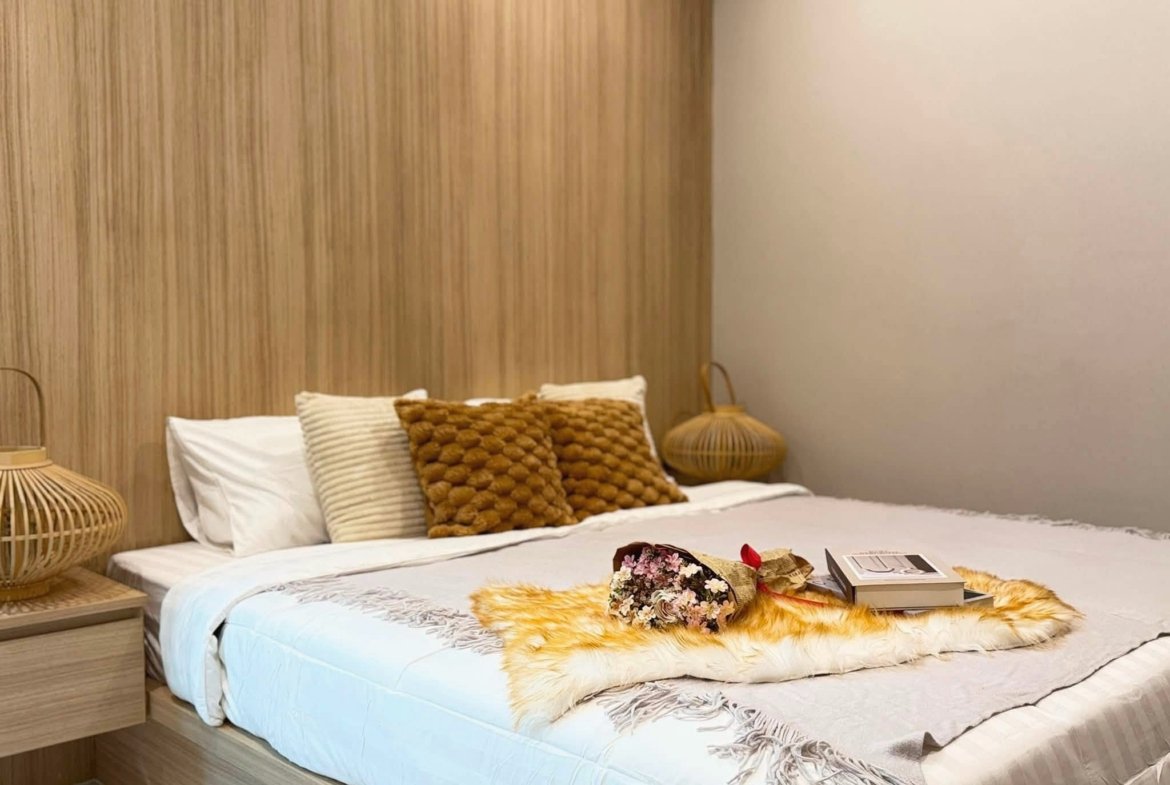Two neatly made twin beds in a cozy bedroom with beige pillows, a wooden paneled wall, and wicker lamps on each side.