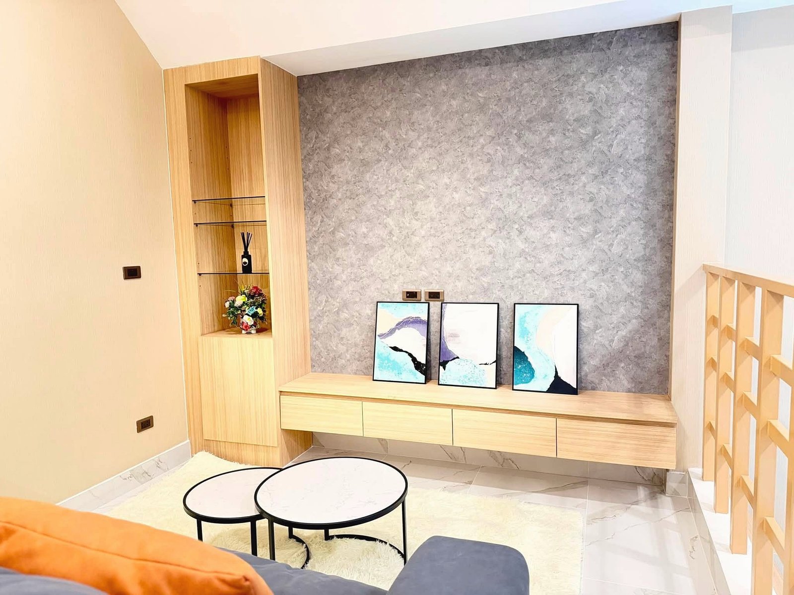 Narita_Villa_Pattaya_Rent_Rdl339_8 Modern living area with light wood built-in shelves, gray textured wall, and framed abstract paintings leaning on a console.